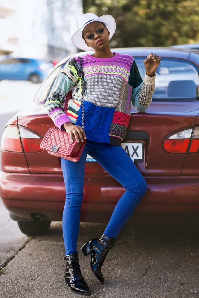 Stylish woman in colorful attire leans against a car in a cityscape background, showcasing modern fashion.