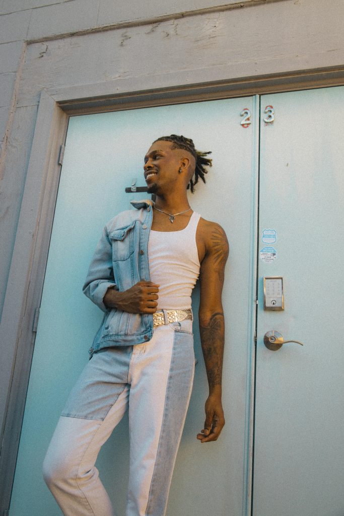 A fashionable man in denim leans against a light blue door, exuding confidence and style.