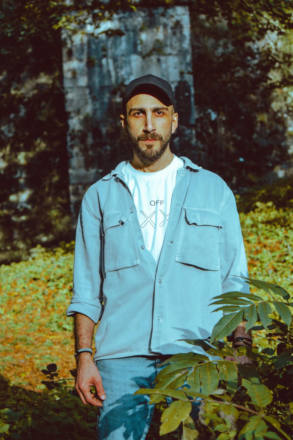 Casual portrait of a man in denim outdoors, surrounded by greenery with a serious expression.
