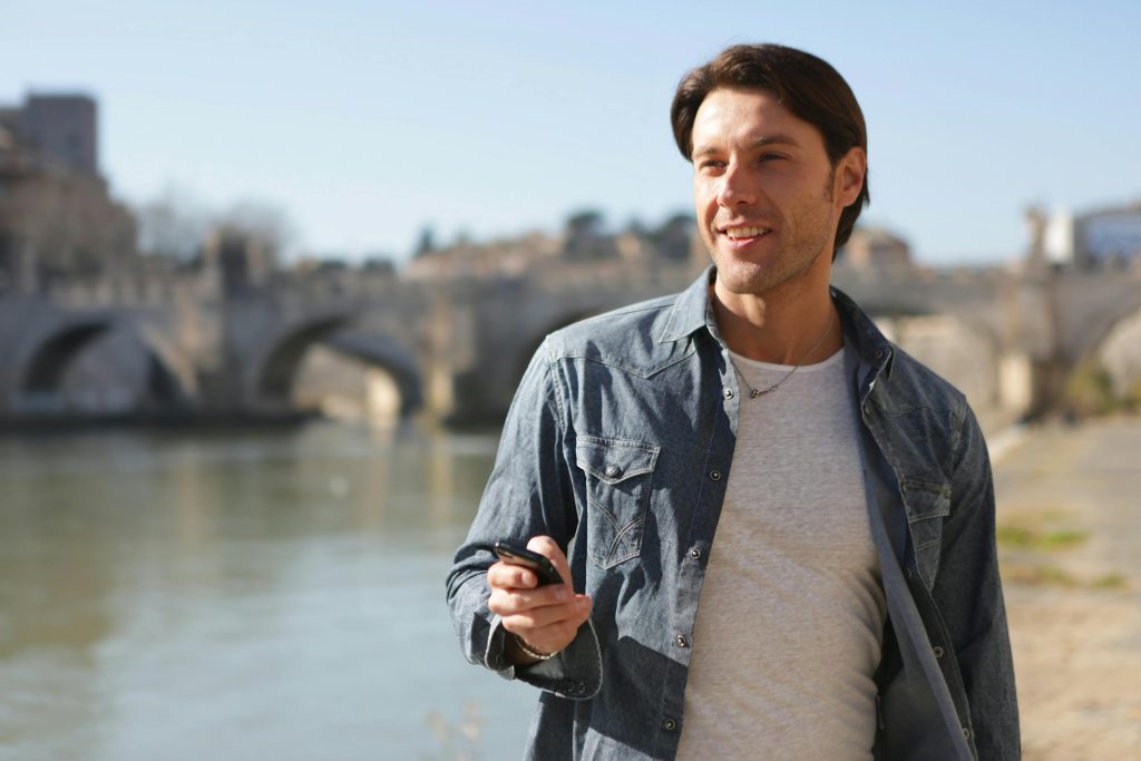 A man enjoys a sunny day by an urban river with a scenic bridge in the background.