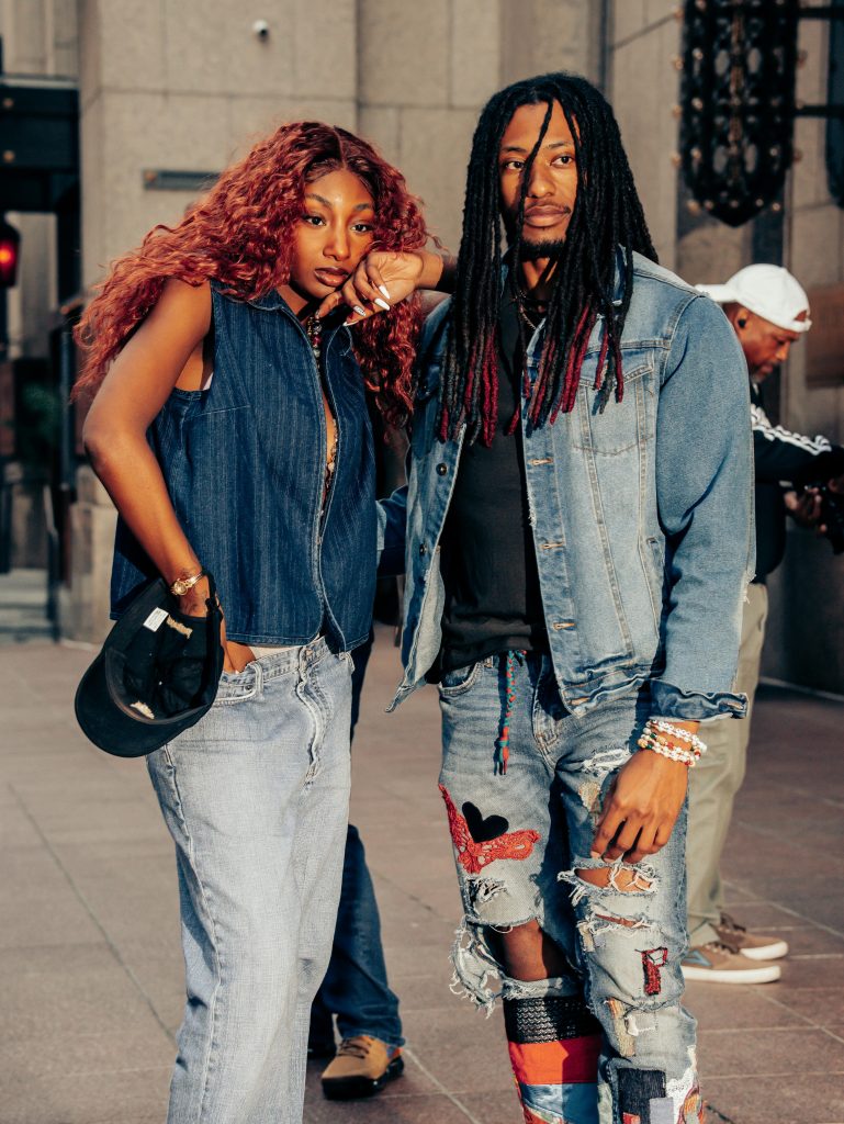 Two adults showcase urban street fashion with denim and dreadlocks in a cityscape.